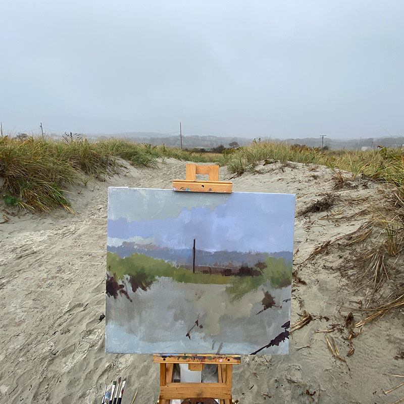 A painting on an easel that is set on a path next to sand dunes near the ocean.