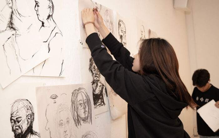 A teenage girl hangs portrait sketches on a wall.