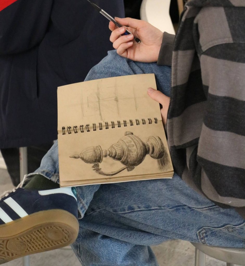 IMG_0281 Student holding a tan sketchbook and pencil, wearing blue jeans, grey and charcoal striped sweater, and navy adidas sambas.